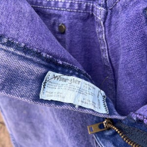 Puede incluir: Primer plano de unos vaqueros Wrangler morados. Los vaqueros tienen cremallera y cierre de bot&oacute;n. Una etiqueta dentro de los vaqueros dice "Wrangler" e incluye instrucciones de lavado. Los vaqueros son 100% algod&oacute;n.
