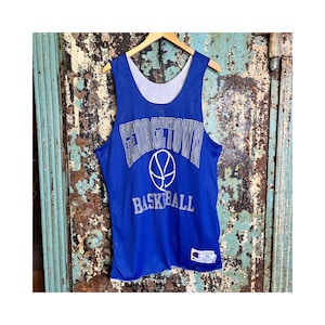 May include: A blue Georgetown basketball jersey with white lettering and a basketball graphic. The jersey is XL and has a white interior. The jersey is hanging on a wooden hanger against a rustic background.