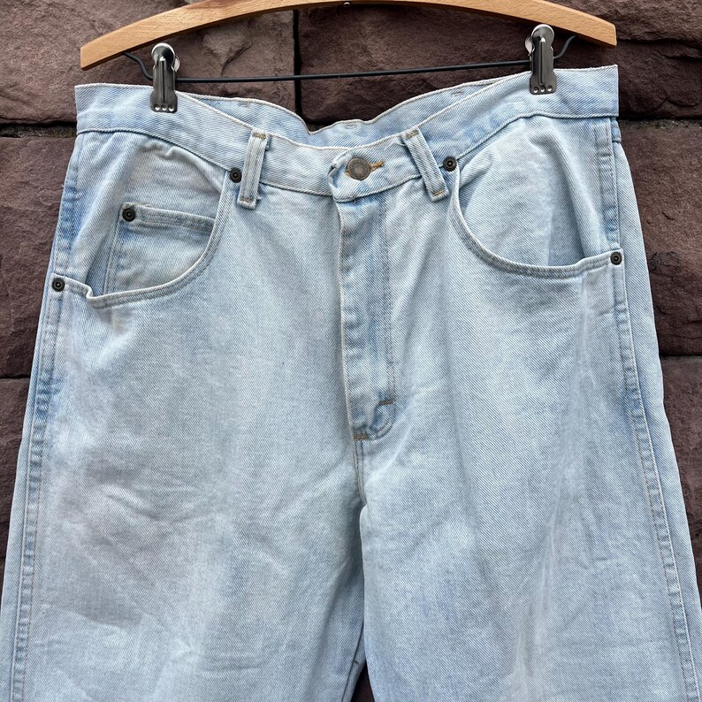 May include: Light blue denim jeans hanging on a wooden hanger. The jeans have a classic five-pocket design, a button and zipper closure, and a straight leg fit. The denim has a faded, worn look.