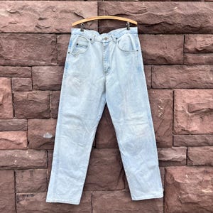 May include: Light blue denim jeans hanging on a wooden hanger. The jeans have a classic five-pocket design and a straight leg cut. The denim has a faded wash, giving it a vintage look. The jeans are displayed against a textured brick wall.