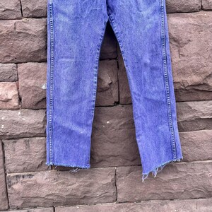 Puede incluir: Vaqueros azules descoloridos con dobladillo sin rematar. Los pantalones vaqueros tienen un corte de pierna recta y se muestran contra una pared de ladrillos texturizada. Los vaqueros tienen un dise&ntilde;o cl&aacute;sico de cinco bolsillos y cierre de bot&oacute;n y cremallera.