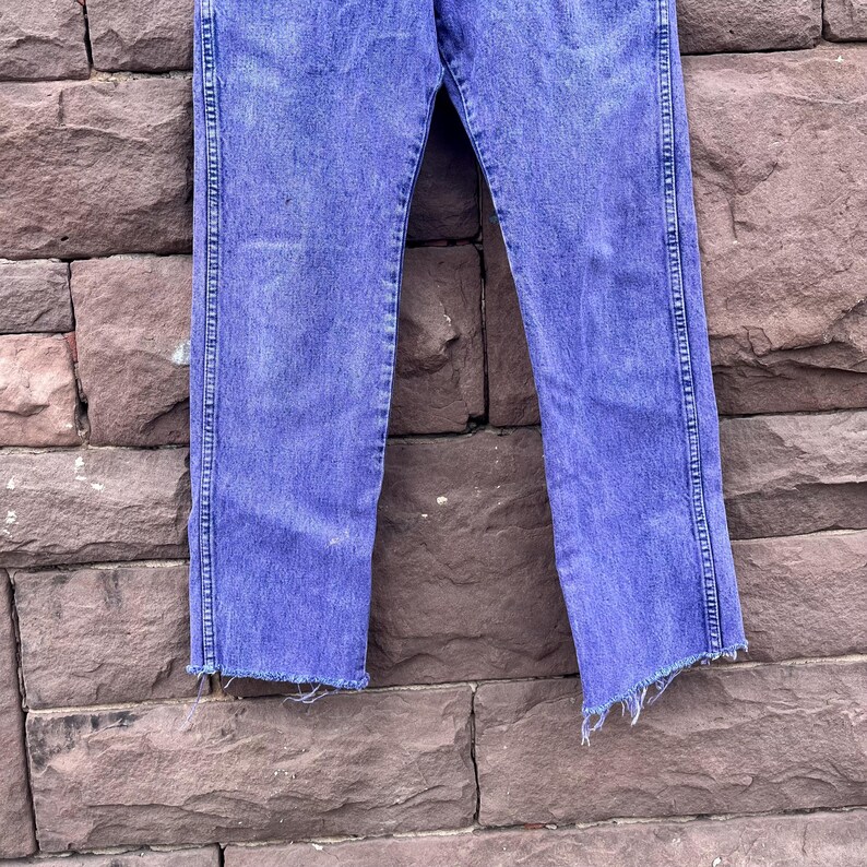 Puede incluir: Vaqueros azules descoloridos con dobladillo sin rematar. Los pantalones vaqueros tienen un corte de pierna recta y se muestran contra una pared de ladrillos texturizada. Los vaqueros tienen un dise&ntilde;o cl&aacute;sico de cinco bolsillos y cierre de bot&oacute;n y cremallera.