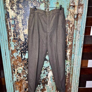May include: Brown trousers hanging on a metal hanger. The pants have a classic design with a button and zipper closure, belt loops, and front pockets. The fabric appears to be a textured wool blend. The trousers are cuffed at the bottom.
