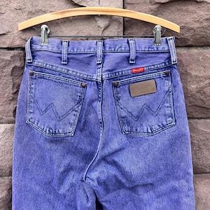 Puede incluir: Un par de vaqueros Wrangler morados vintage, colgados de una percha de madera. Los vaqueros presentan el logotipo cl&aacute;sico de Wrangler en el bolsillo trasero y una etiqueta roja con el logotipo. La mezclilla tiene un aspecto descolorido y desgastado.