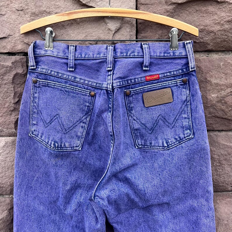 Puede incluir: Un par de vaqueros Wrangler morados vintage, colgados de una percha de madera. Los vaqueros presentan el logotipo cl&aacute;sico de Wrangler en el bolsillo trasero y una etiqueta roja con el logotipo. La mezclilla tiene un aspecto descolorido y desgastado.