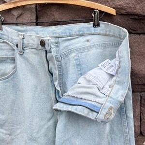 May include: Light blue denim jeans with a button and zipper closure. The jeans are hanging on a wooden hanger. The inside of the jeans is visible, showing the label with the size 34 x 32.