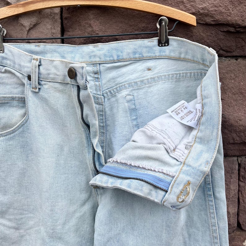 May include: Light blue denim jeans with a button and zipper closure. The jeans are hanging on a wooden hanger. The inside of the jeans is visible, showing the label with the size 34 x 32.