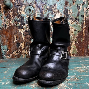 May include: A pair of worn black leather motorcycle boots with buckle straps. The boots have a rounded toe and a short shaft. The background is a distressed, weathered door with a turquoise and brown color scheme.