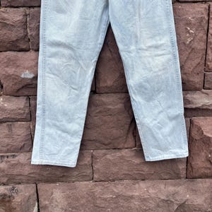 May include: Light blue denim jeans. The jeans are made of a light wash denim material. The jeans have a classic five-pocket design and a straight leg fit. The jeans are displayed against a brick wall.
