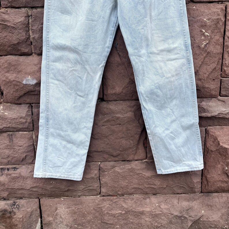 May include: Light blue denim jeans. The jeans are made of a light wash denim material. The jeans have a classic five-pocket design and a straight leg fit. The jeans are displayed against a brick wall.