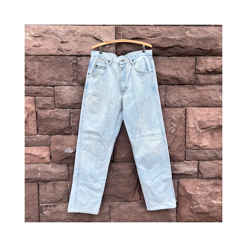 May include: Light blue denim jeans hanging on a wooden hanger against a brick wall. The jeans have a classic five-pocket design and a straight leg cut. The denim has a faded, worn appearance.