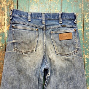 Pode incluir: Jeans de ganga azul desbotada com o log&oacute;tipo Wrangler num remendo de couro castanho. Os jeans t&ecirc;m dois bolsos traseiros com o log&oacute;tipo Wrangler cosido. Os jeans s&atilde;o vintage e mostram sinais de desgaste.