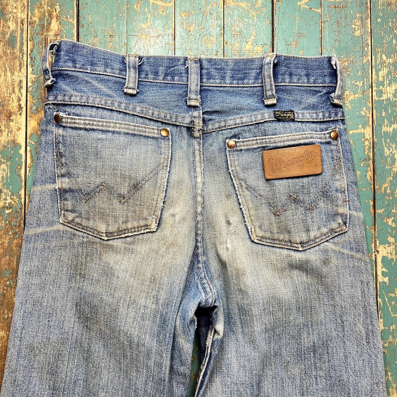 Pode incluir: Jeans de ganga azul desbotada com o log&oacute;tipo Wrangler num remendo de couro castanho. Os jeans t&ecirc;m dois bolsos traseiros com o log&oacute;tipo Wrangler cosido. Os jeans s&atilde;o vintage e mostram sinais de desgaste.