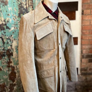 May include: A tan suede jacket with four front pockets and a button-up closure. The jacket has a wide collar and is displayed on a mannequin. A red and black plaid shirt is visible underneath the jacket.