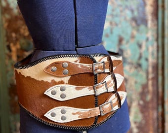 May include: A brown and white cowhide belt with three leather straps and silver buckles. The belt is on a blue mannequin and has black stitching around the edges. The belt is a wide style.