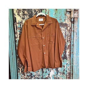 May include: A long-sleeved, rust-colored shirt with two chest pockets and white buttons. The shirt is hanging on a white hanger against a distressed, turquoise and brown background. The shirt has a classic collar and buttoned cuffs.