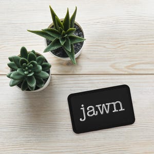 May include: A rectangular black embroidered patch featuring the white lowercase text jawn in a serif font, positioned alongside two small green succulent plants in white pots, showcasing a clean and simple aesthetic arrangement.