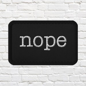 May include: A rectangular black embroidered patch with rounded corners featuring the word nope stitched in white thread in a lowercase serif font. The patch has a thick black stitched border around the entire perimeter of the fabric rectangle.