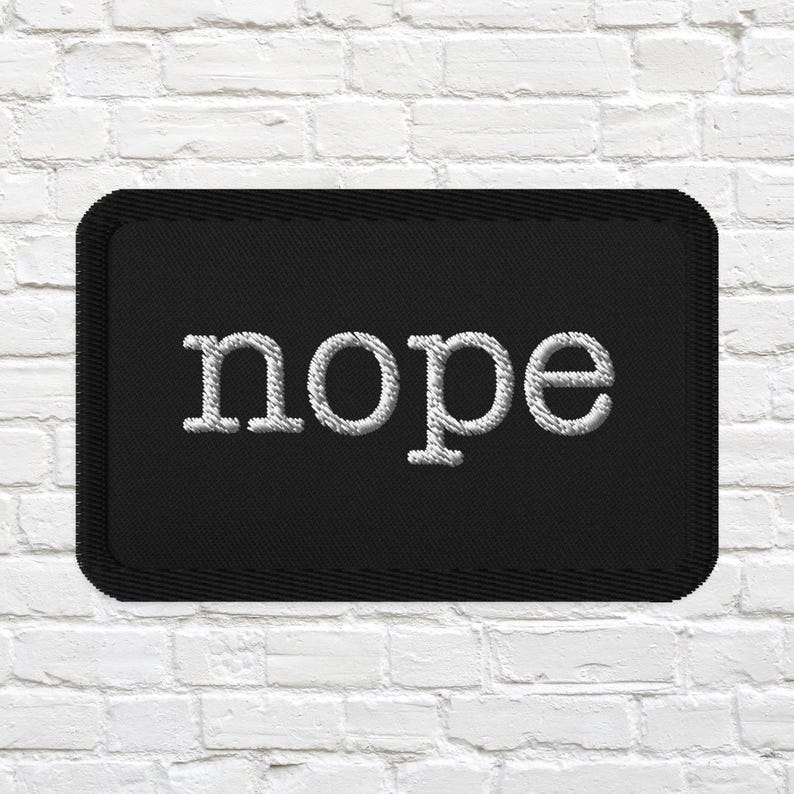 May include: A rectangular black embroidered patch with rounded corners featuring the word nope stitched in white thread in a lowercase serif font. The patch has a thick black stitched border around the entire perimeter of the fabric rectangle.