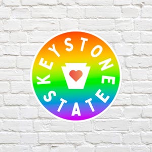 Puede incluir: Una pegatina circular con un fondo de degradado de arcoíris vibrante. El texto KEYSTONE STATE está impreso en letras mayúsculas blancas y negritas alrededor del centro. Un contorno blanco del estado de Pensilvania con un corazón rojo.