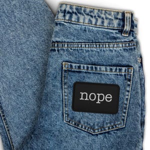May include: Acid-wash blue denim jeans featuring a high-waisted design with belt loops and a single back pocket. A rectangular black patch is stitched onto the back pocket, displaying the word nope in white, lowercase, serif-style lettering.