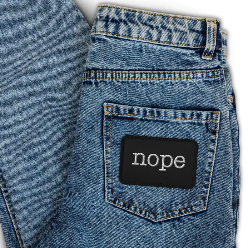 May include: Acid-wash blue denim jeans featuring a high-waisted design with belt loops and a single back pocket. A rectangular black patch is stitched onto the back pocket, displaying the word nope in white, lowercase, serif-style lettering.