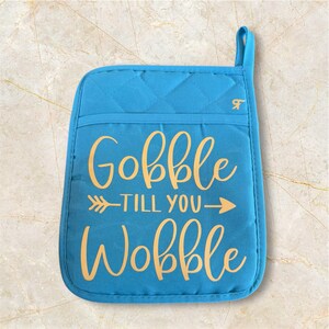 May include: Blue quilted oven mitt with a gold inscription that reads "Gobble 'til you wobble" with an arrow pointing to the right.