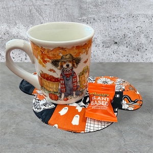 May include: A white ceramic mug featuring a watercolor illustration of a dog in a straw hat and overalls standing near baskets of pumpkins, resting on a quilted fabric coaster with a black and orange pattern of ghosts and moths, with a Reese's candy.
