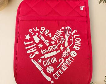 Op de afbeelding: Rode gewatteerde ovenwant met een wit hartvormig ontwerp met bak ingrediënten zoals suiker, bloem en cacao. De want heeft een lus om op te hangen en een zak. Feestelijk keukengerei.