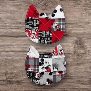 May include: Two cat-shaped quilted pot holders. The pot holders feature a patchwork design with red, black, white, and gray fabrics. The fabric includes images of Mickey and Minnie Mouse, along with text that reads "Minnie All About the Dots".