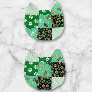 May include: Two cat-shaped quilted coasters. Each coaster is a patchwork of green fabrics with white paw prints, mice, and shamrocks. One section has gold glitter shamrocks. The coasters are designed to protect surfaces from heat and spills.