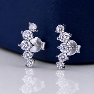May include: A pair of silver-toned stud earrings, each featuring five round, clear, faceted stones arranged in a curved, ascending line. The earrings have a metallic post backing and a polished finish, reflecting light across the circular stones.