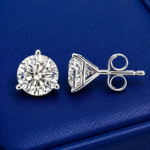 May include: A pair of silver-toned stud earrings featuring round, brilliant-cut clear stones. One earring is shown from the front, displaying the faceted surface, while the other is shown from the side, revealing the three-prong setting and post back.