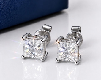 May include: A pair of silver-toned stud earrings featuring square-cut clear stones held in a four-prong setting. The earrings have a reflective metallic finish and are designed with a post back mechanism for secure attachment to the earlobe.