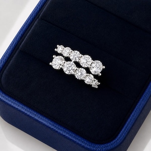 May include: A silver-toned ring featuring two parallel rows of round, clear, faceted stones. The ring is displayed inside a dark blue velvet-lined jewelry box with a textured blue exterior, showcasing a multi-stone design with a polished metal band.