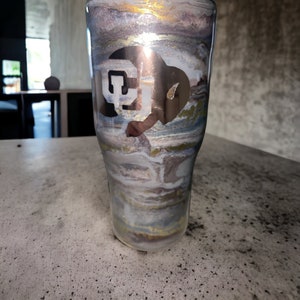 Może przedstawiać: Kubek ze stali nierdzewnej z marmurowym wzorem w odcieniach bieli, szarości i złota. Kubek ma białą naklejkę winylową z literami "CU" w pogrubionym kroju.