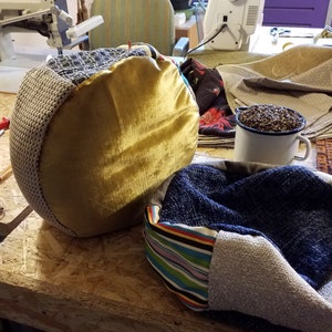 May include: Two handmade fabric ottomans in a workshop setting. One ottoman features a gold-colored top and a gray knitted side. The other has a blue fabric interior and a striped fabric edge. A white mug filled with seeds sits nearby.