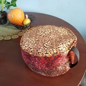 May include: A round, multi-colored, fabric meditation cushion with a black strap handle. The cushion has a top layer of gold and brown fabric with a leaf pattern, a middle layer of red and orange fabric with a leaf pattern, and a bottom layer of brown fabric.