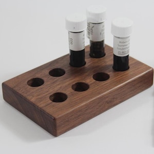 May include: A wooden ink bottle holder with eight slots, three of which are filled with ink bottles. The holder is made of dark wood and has a rectangular shape. A purple fountain pen is lying on the table next to the holder.