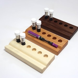 May include: Three wooden ink bottle holders, each with a different wood finish: light wood, medium wood, and dark wood. Each holder has six round holes for ink bottles and a groove for a pen. The holders are stacked on top of each other, with the light wood holder on the bottom and the dark wood holder on top. A purple pen is resting in the groove of the medium wood holder.