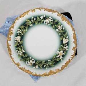 May include: A white porcelain plate with a gold scalloped edge. The plate features a floral wreath design with white flowers and green leaves against a gradient green background. The plate is resting on a blue cloth.