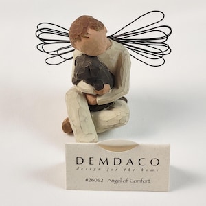 May include: A handcrafted figurine of an angel, kneeling and embracing a dark-colored animal. The angel has brown hair and wears a cream-colored robe. Black wire wings extend from the back. A small card reads "Angel of Comfort."