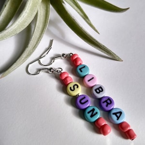 May include: A pair of colourful beaded earrings spelling out "SUN" and "LIBRA". The beads are in shades of pink, yellow, purple, blue and green.