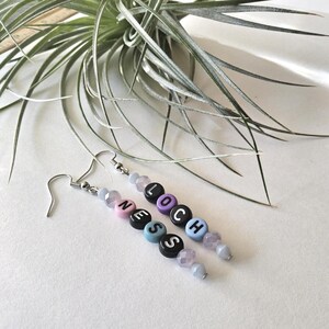 May include: A pair of handmade earrings with the word "LOCH NESS" spelled out in colorful letter beads. The earrings feature silver-colored hooks and are accented with light purple and blue beads. A sprig of green foliage is in the background.