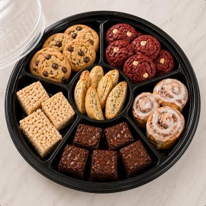 May include: A black, divided serving tray filled with an assortment of baked goods. Chocolate chip cookies, red velvet cookies, brownies, biscotti, rice crispy treats, and cinnamon rolls are arranged in separate sections.