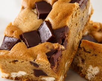 May include: A thick, golden-brown cookie bar featuring large, square chunks of dark chocolate embedded throughout the dough. The baked treat has a soft, dense texture with visible white chocolate chips mixed into the base of the cookie dough.