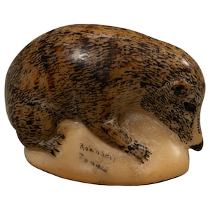 May include: A carved stone figurine of a bear in a crouching position, featuring a mottled brown and black textured surface. The base is a smooth, light cream color with the handwritten text Robinson Inukjuak visible on the front side.