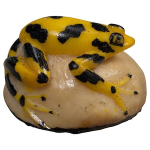 Pode incluir: Uma pequena estatueta de sapo com pele amarelo-brilhante e manchas pretas irregulares, repousando sobre uma base lisa, arredondada e de cor esbranquiçada que parece uma pedra. O sapo está numa posição agachada com os membros contra a superfície pálida.