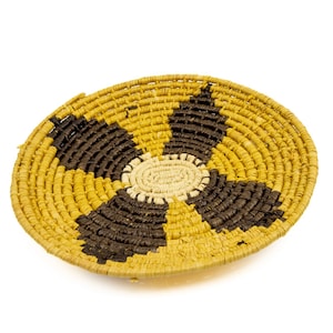 May include: A round, woven basket with a floral design. The basket features a yellow and brown color scheme, with a central cream-colored circle. The design is symmetrical, with the brown sections forming a flower shape.