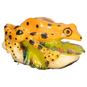 May include: A small, yellow frog with black spots is sitting on a green and brown leaf. The frog is made of ceramic and has a realistic appearance.